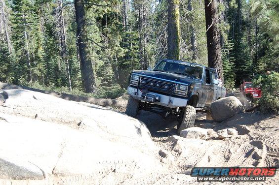 Dark blue Ford pickup truck with aftermarket bumper and winch navigating a rocky trail, red Jeep visible in background