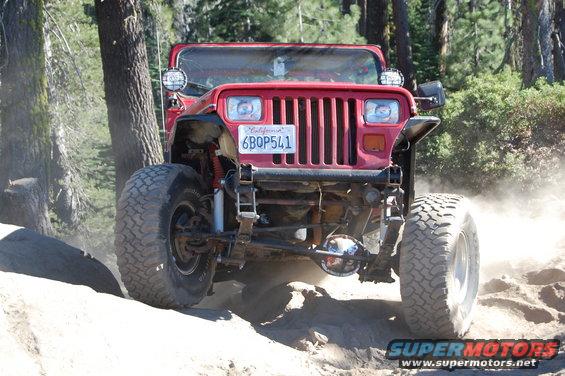 Red 1993 Jeep Wrangler driving on a dirt trail, kicking up dust. It has large off-road tires and auxiliary lights