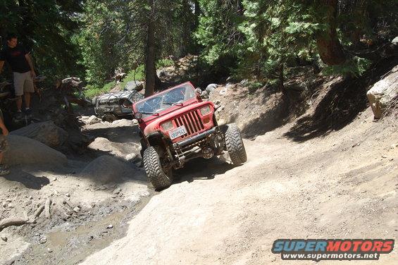 Red Jeep Wrangler on a dirt trail, front grille visible, license plate reads 6BDP643. Another vehicle is behind it
