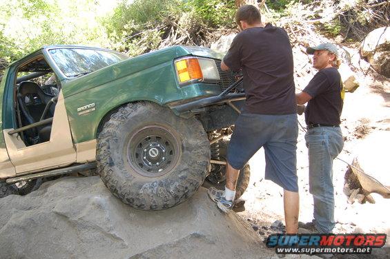 Green and tan off-road vehicle with roll cage, large tires, and aftermarket bumper on rocky terrain. Two people are near the front