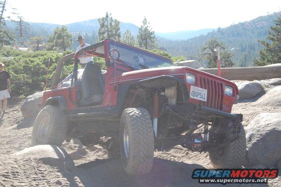 Red Jeep Wrangler on rocky terrain, driver in hat visible