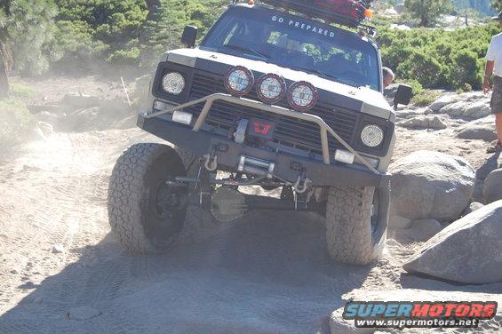 Modified off-road vehicle with winch and extra lights navigating a rocky trail, kicking up dust