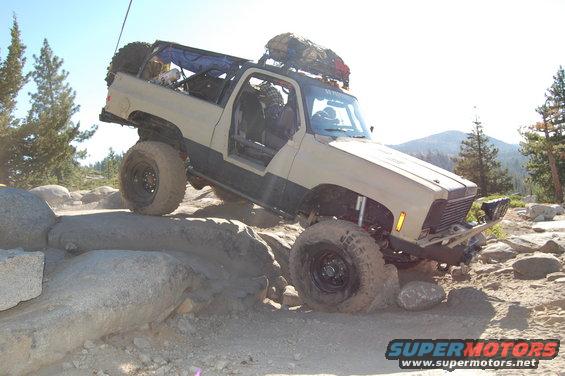 Tan off-road SUV with large tires navigating rocks, doors removed, spare tire on back, roof rack with gear