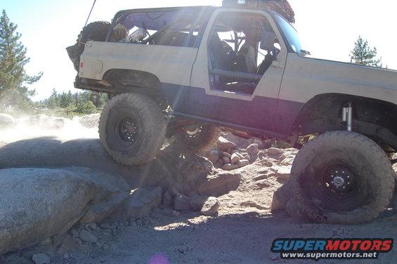 Off-road vehicle with large tires navigating a rocky terrain. The vehicle has a roll cage and is partially covered