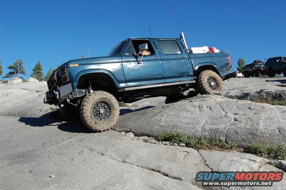 Blue pickup truck with off-road tires climbing rocks, aftermarket bumper, driver visible