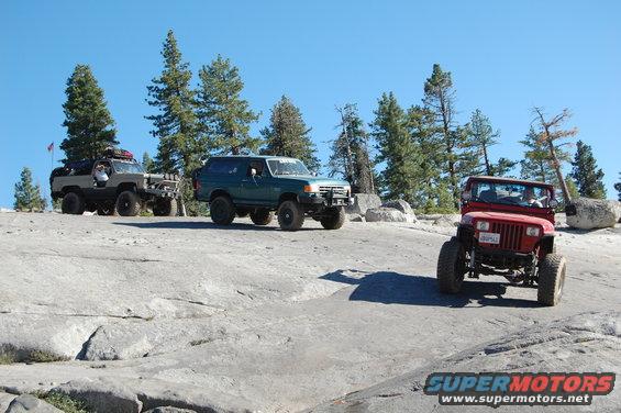 Red 1993 Jeep Wrangler driving on rocks, with a green SUV and a grey truck behind it