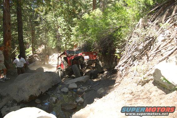 Two red Jeep Wranglers navigating a difficult, rocky off-road trail with trees and brush on either side. One Jeep is in the foreground, the other behind it