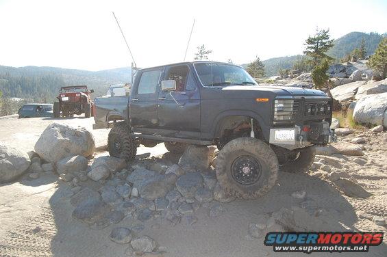 Lifted dark blue Ford F-Series crew cab truck with large tires on rocky terrain. Another red Jeep and a blue van are visible in the background