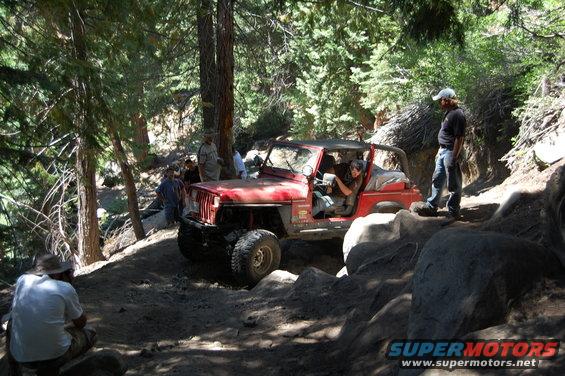 Red 1993 Jeep Wrangler navigating a rocky, wooded trail with people watching