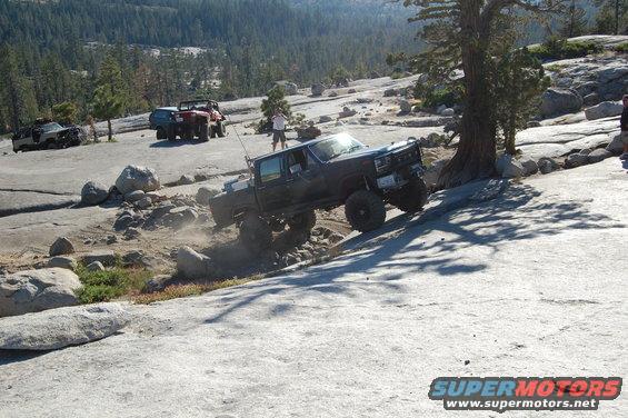 Dark pickup truck with large tires climbing a rocky incline, front-right wheel elevated