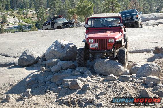 Red 1993 Jeep Wrangler navigating rocks on a trail, with another Jeep and a van behind it