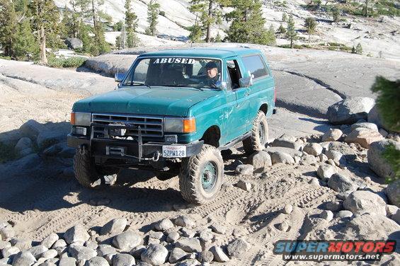 Green Ford Bronco with "ABUSED" sticker on windshield, off-roading on rocky terrain