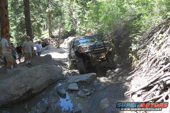 Off-road vehicle navigating a rocky, wooded trail with people watching