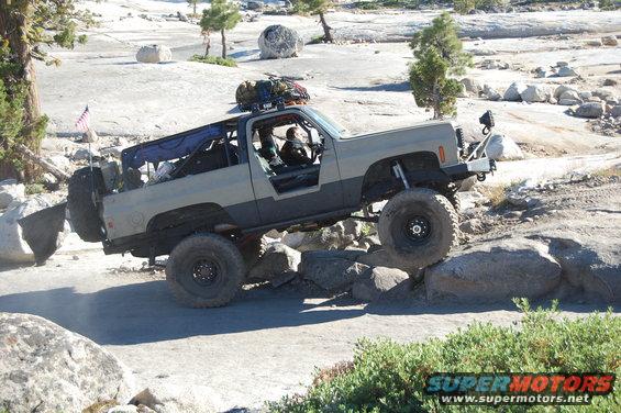 Tan and grey off-road SUV with large tires, driving over rocks. An American flag is mounted on the rear