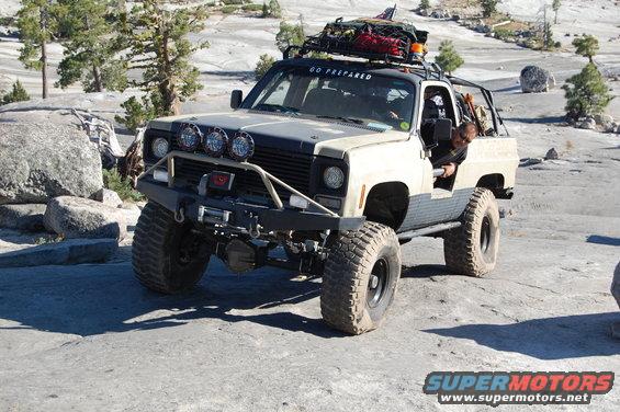 Tan and grey off-road truck with large tires and roof rack, parked on rocky terrain