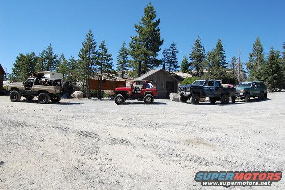 Four off-road vehicles parked in a dirt lot with trees and buildings in the background