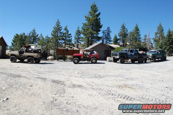 Four off-road vehicles parked in a dirt lot with trees and a cabin in the background