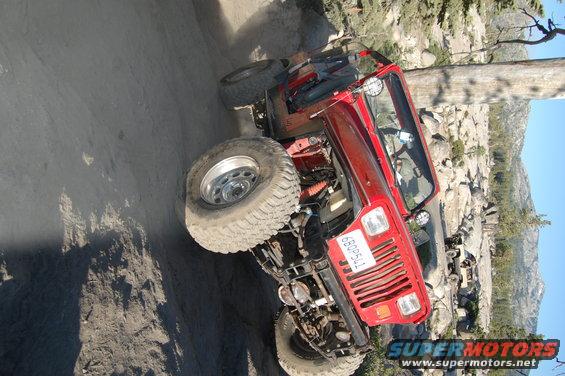 Red 1993 Jeep Wrangler on a rocky trail, front grille and tires visible. Another vehicle is parked in the background