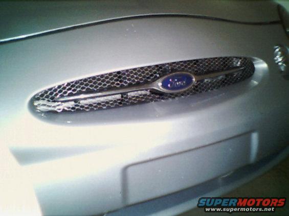 Silver Ford Taurus grille with blue oval Ford badge and honeycomb mesh. Visible portion of bumper below