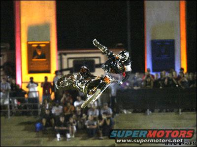 Motocross rider mid-air performing a stunt on a dirt bike, with spectators in the background