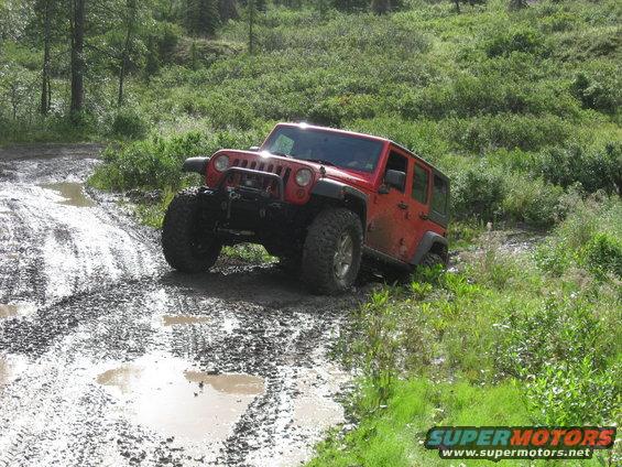 Red Jeep Wrangler driving through mud and puddles on a dirt path with green foliage on the sides