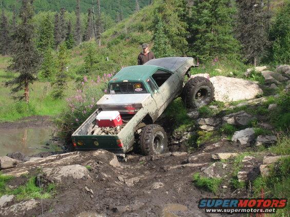 Mud-covered green and tan pickup truck with large tires climbing a rocky, muddy hill