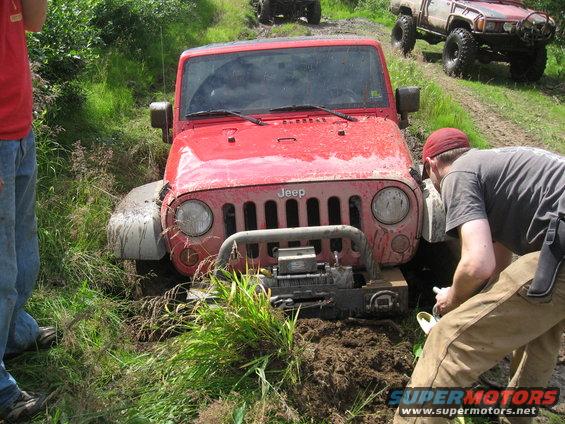 Red Jeep stuck in mud, people trying to free it. Another truck is visible in the background