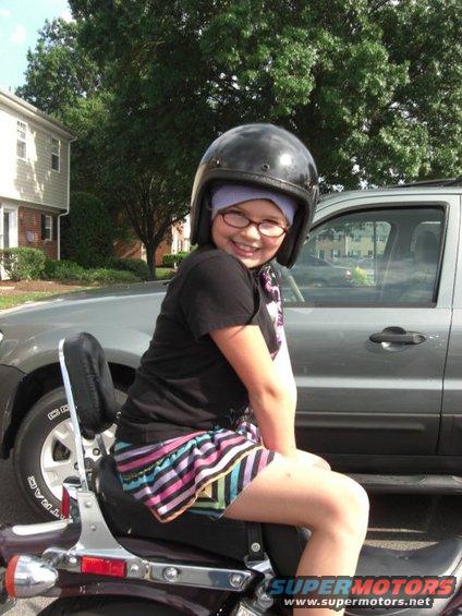 Girl in helmet and striped skirt sits on a motorcycle, with a silver SUV behind her
