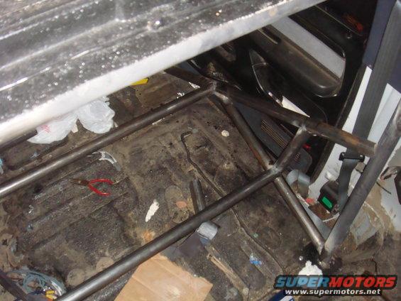 Interior view of a vehicle with a metal roll cage installed, showing a dirty floor and seatbelt