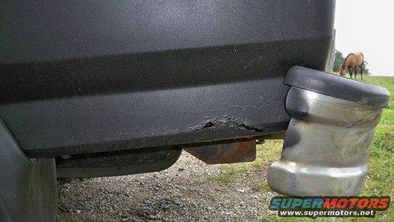 Close-up of rusted rear bumper corner on a dark-colored SUV, with a horse grazing in the background