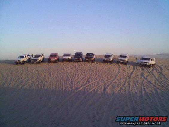 Line of SUVs parked on sand dunes under a clear sky. One white SUV has its door open