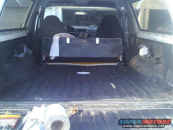 Interior cargo area of a light-colored SUV, showing black seats and a dark floor liner. A roll of material and scissors are on the tailgate
