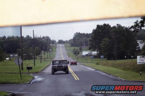 Humvee and red car on a two-lane road with trees and buildings