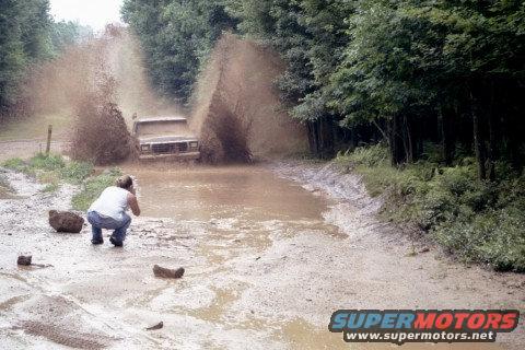Truck driving through a muddy puddle, kicking up a lot of water and dirt. A person is crouching down to take a photo