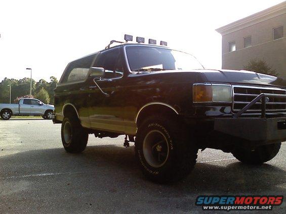 Black 1990 Ford Bronco with roof light bar and custom front bumper parked next to a silver pickup truck