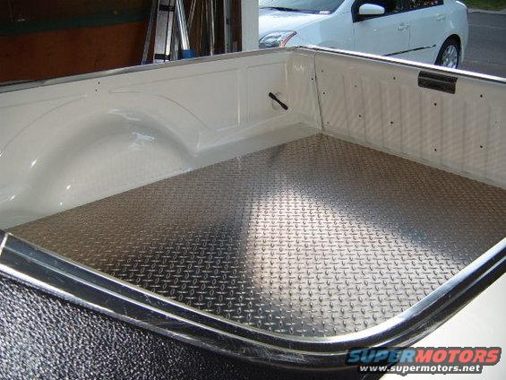 Truck bed with diamond plate flooring and white exterior. A white sedan is visible in the background