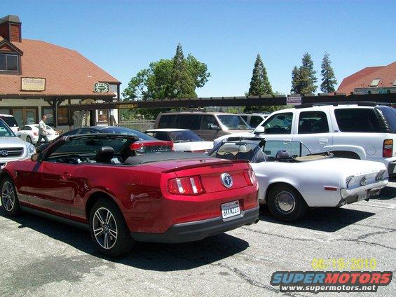 Red convertible Mustang parked next to a white classic convertible Mustang. A white SUV is behind them