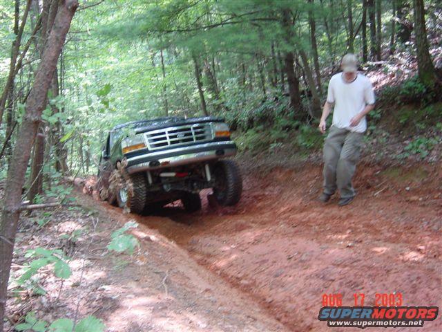 Ford Bronco stuck in mud on a wooded trail. Guy in white shirt and cargo pants walks nearby