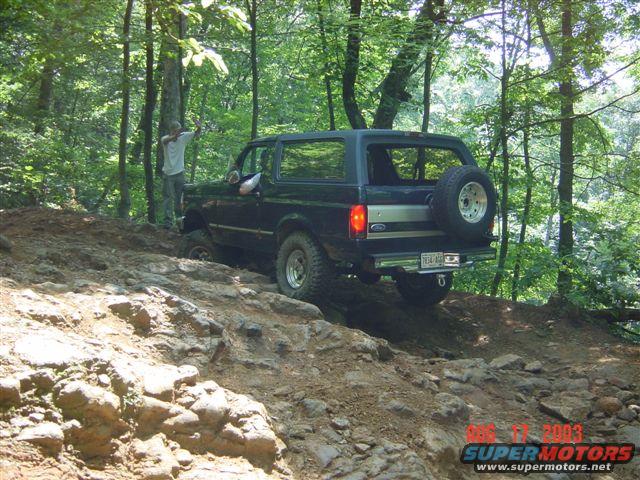 Black 1995 Ford Bronco climbing a steep, rocky trail in a wooded area. A person stands by the driver's door