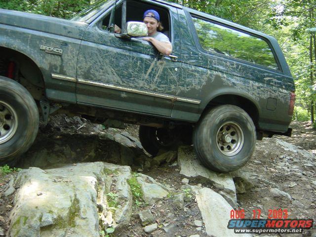 Green 1995 Ford Bronco driving over rocks on a wooded trail. Driver is wearing a blue baseball cap
