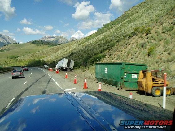Roadside accident scene with overturned trailer, green dumpster, and yellow skid steer loader. Cars drive past on the highway