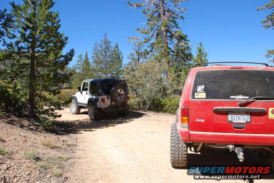 Red Jeep Cherokee and white Jeep Wrangler on a dirt road with trees and blue sky