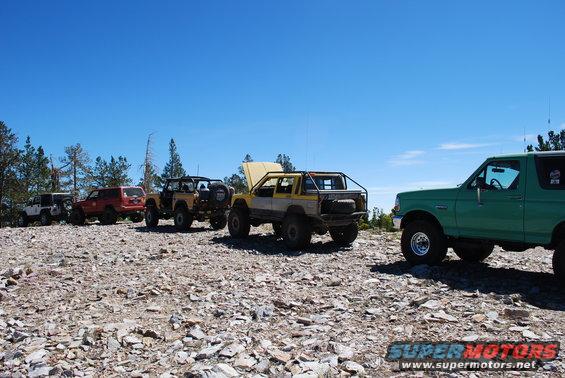 Several off-road vehicles parked on a rocky hill under a clear blue sky. One green Bronco is in the foreground