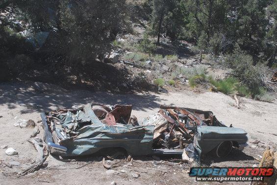 Wrecked green car from the side, heavily damaged with exposed interior and rusted parts. Visible chrome bumper and wheel