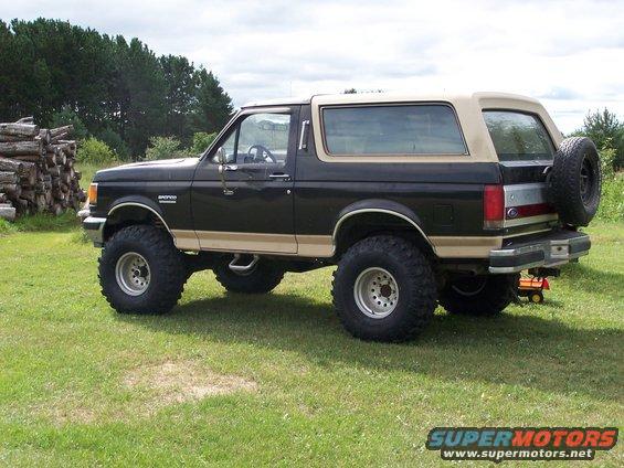 Lifted black and tan Ford Bronco with large tires parked on grass, pile of logs to the left