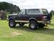 1987 Ford Bronco - This lifted Ford Bronco is parked on a grassy field with a pile of logs nearby.