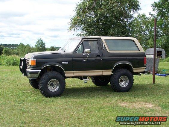 Black and tan 1987 Ford Bronco with large tires and a brush guard, parked on grass