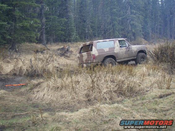 Camouflaged 1985 Ford Bronco driving through a muddy, grassy area with trees in the background