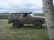 1985 Ford Bronco - A muddy SUV, likely from the 1980s, sits off-road near a large tree under an overcast sky.