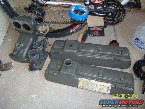 Gray engine intake manifold and valve covers on concrete floor with bicycle wheel and tape